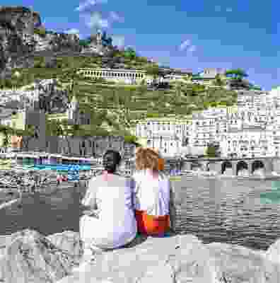 Two women sat on the edge of a rock out at sea, looking at Positano's beach shore.