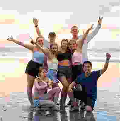 Group of travellers posing with their arms in the air on the beach shore at sunset.