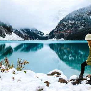 A person standing on snow in front of a bright blue lake and pine forested mountains